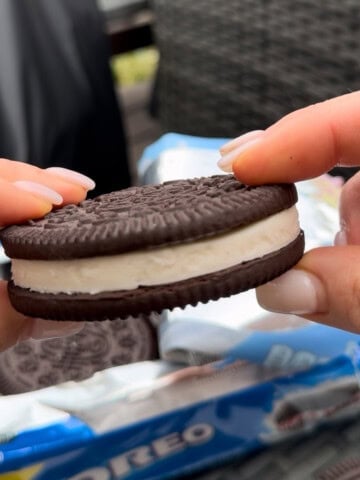 Hand holding a huge Oreo cookie with thick cream filling beside a package of Oreos, showing the size and texture up close.
