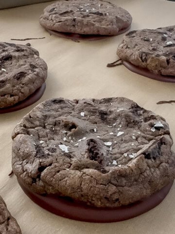 Close-up of bakery-style cookies and cream cookies made from Pillsbury Oreo cookie dough, topped with flake salt and dipped in melted chocolate on parchment paper.